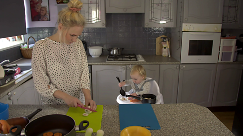 Woman chopping mushrooms in kitchen while her toddler sits in a high chair