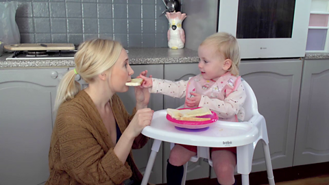 Woman eating toast handed to her by toddler in high chair.