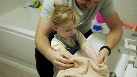 Man sat on side of bath next to sink using towel to dry hands of toddler