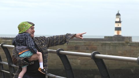 Man carrying a little boy, looking out to a lighthouse. 