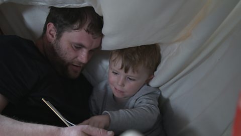 Little boy and dad under a blanket reading a book.