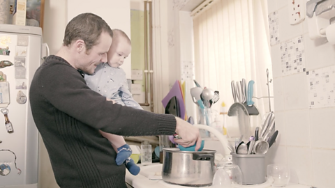 Dad carrying baby whilst washing up dishes. 