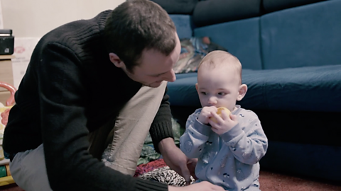 Dad looking at baby, who has got a toy in his mouth.