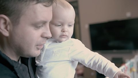 Dad holding baby, baby is pointing and looking down. 