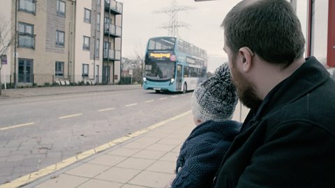 Baby and dad at a bus stop looking at a bus.