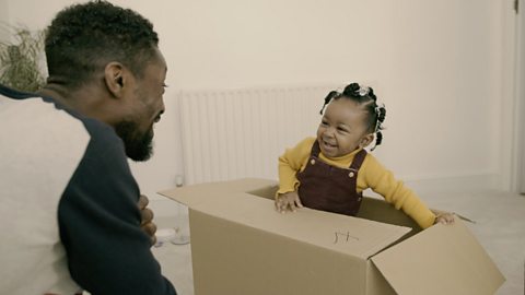 Dad watching daughter sat in a cardboard box.