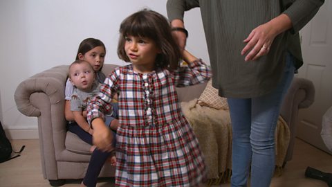 Girl in checked dress holding hands with mum while her family look on from sofa.