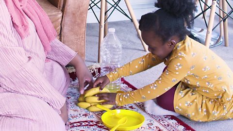 Girl and mum sat on rug playing with yellow plate, yellow cutlery and bananas.