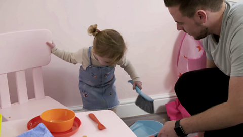 Little girl holding on to chair and crouching down to use a dustpan and brush