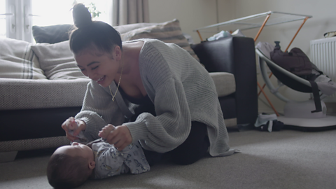 Mum crouched down and holding hands of her baby who is lying on the floor