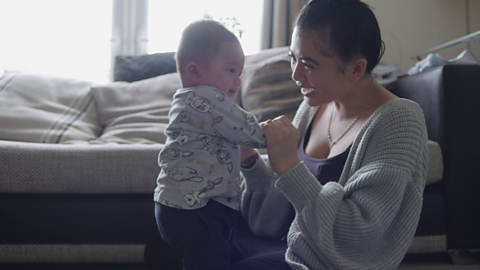 Mum holding hands of her baby who is stood in her lap.