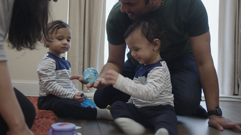 Two toddlers sat on floor playing while parents crouch next to them.