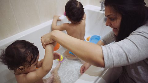 Mum holding the hand of her toddler sat in bath as she kneels by bathtub.