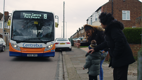 Family of three stood at bus stop and waving at approaching bus.