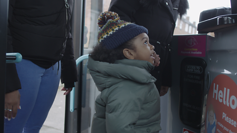 Child wearing bobble hat and talking to bus driver.