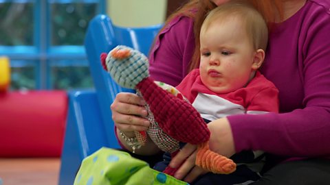 Baby sat on his mum's knee, looking at a soft toy.