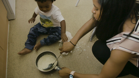 Toddler sat on kitchen floor with mum who is stirring a pan with spoon.