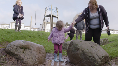 Toddler holding hand of mum and standing in a puddle.