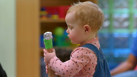 A baby in a pink and blue dress plays with a shaker filled with porridge oats.