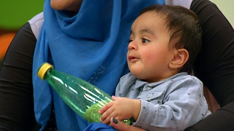 A young baby sits on his mother's lap with a porridge oat rattle.