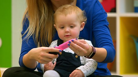 A small baby sits on her mum's lap and looks at a small, pink hairbrush mirror.