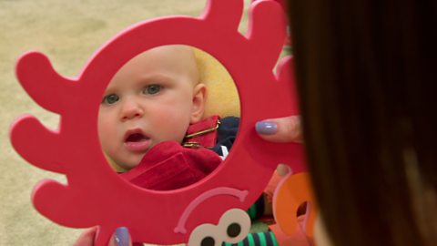 Baby in dungarees looks unimpressed at his reflection.