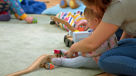 Young baby and her mother play with a toy train on a track