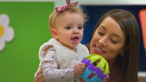 A baby stands with her mum, holding a shaker toy