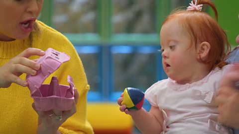 A baby holding a juggling ball looking at an open egg carton.