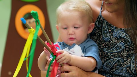 A young baby lies on the floor, reaching for sticks in his mum's hand.