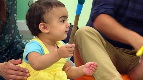 A baby in a yellow dress plays with a stick at the Baby Club