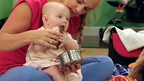   A young baby puts a small, metal, pan in their mouth.