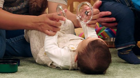 A young baby lies on the floor with pot lids in both hands.