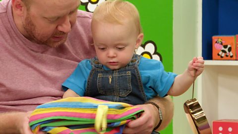 A small baby pulls a toy frying pan out of the Baby Club bag.