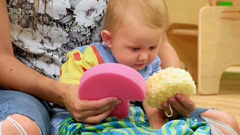 A baby chooses between two sponges being offered to him.