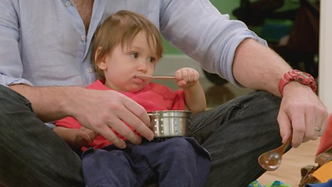 A baby sitting in a parent's lap with a spoon in their mouth