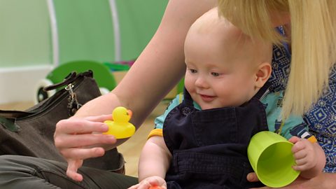 A parent with their baby holding a yellow rubber duck and a green plastic cup