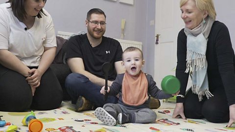 A mum, dad and health visitor gathered around a baby boy hitting a pan with a spoon.