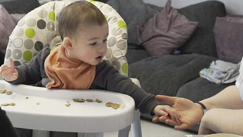 A little boy in a high chair reaching out to his mum's hand.