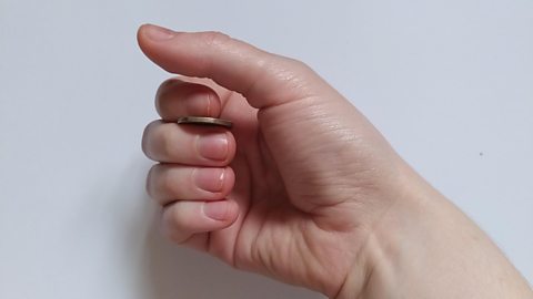 Close up of a hand holding a 20p piece between the index and middle finger, the fingers curled towards the thumb.