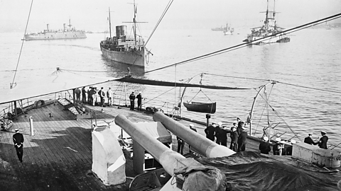 A photograph taken from onboard a battleship of World War One with naval officers looking out to sea with five naval ships at rest.