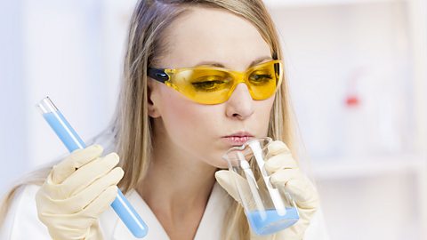 A woman wearing a white lab coat and safety goggles sniffing a beaker containing a blue substance.