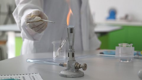 A person in a lab coat and gloves holding an object close to the flame of a lit bunsen burner.