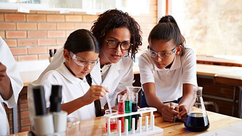 Teacher assisting two students with science project involving test tubes and a conical flask.