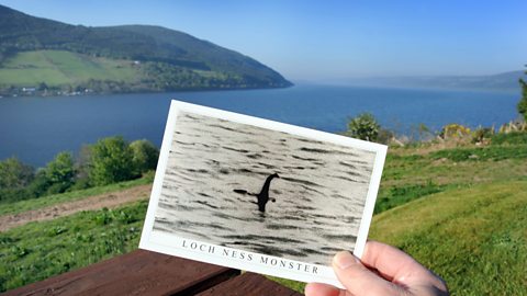 A hand holding a postcard of a black and white photograph with the infamous Loch Ness monster sighting in front of Loch Ness 