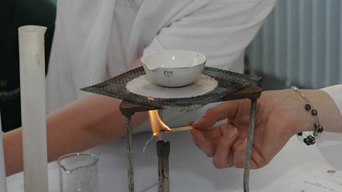 A Bunsen burner is beneath a tripod and gauze, to turn a liquid into a gas. A hand is lighting the Bunsen burner.