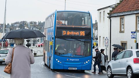 A double decker bus heading to St Andrews. There are pedestrians with umbrellas up walking by the bus.