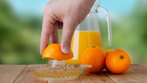 A hand juices an orange using a glass juicer. Oranges and jug in background