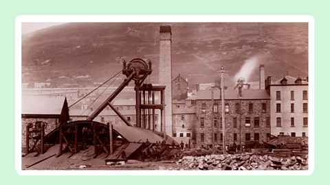 A tall metal structure, crowned with a wheel towing coal from out of a mine at the back of a row of terraced houses.