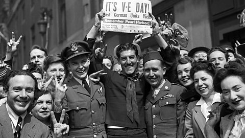 A crowd of people celebrating VE Day and holding up a newspaper with the headline 'it's VE Day!.' 